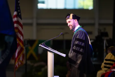 A bearded professor in cap and gown speaks to audience at commencement.