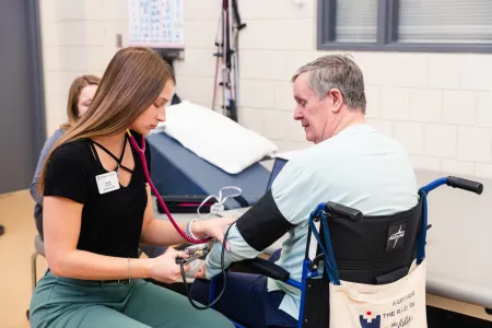 Student supporting an elderly man in his medical routine, led by a Professor of Aging Studies at Ithaca College