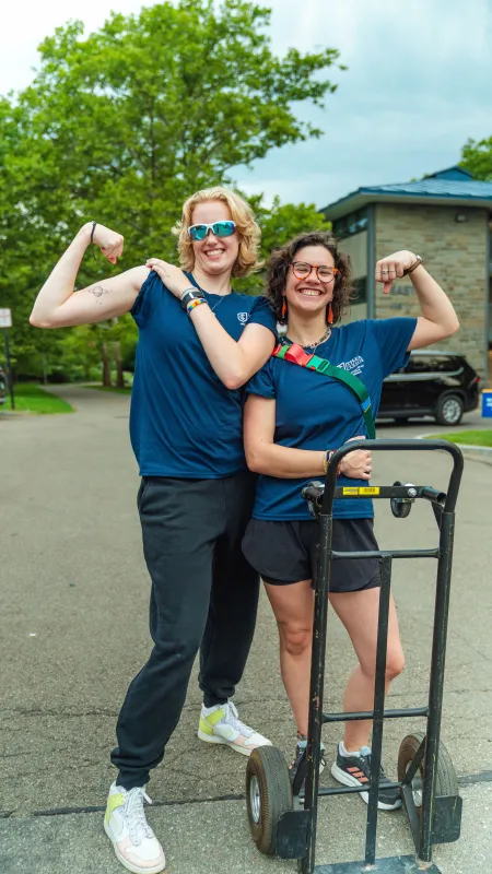 Two college students pose with a hand truck and flex their biceps.