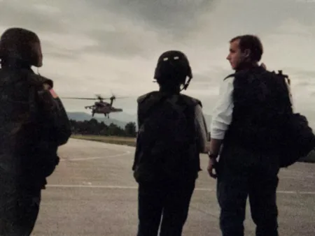 Three men, two in military gear, one in civilian gear, at an airstrip.