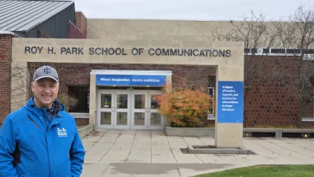 A man stands on a college campus in front of a building labeled "Roy H. Park School of Communications."
