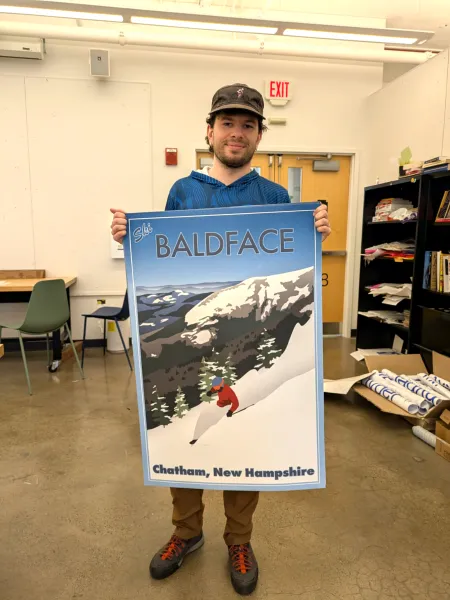 Adams Staples holds a large travel-style poster he designed, showing a skier in a red jacket on a snowy slope with the text "Ski Baldface, Chatham, New Hampshire."