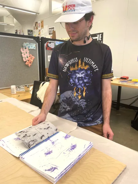 Adams Staples, in a Lobster Pot 2024 cap, turns the pages of a spiral-bound book of abstract purple and black prints in a design studio.