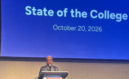 A woman speaks at a podium in front of a projection that reads "State of the College, October 20, 2026."