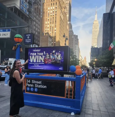Ithaca College Alumni Maya Rodgers '20 points to a "For the Win" ad above the 34 Street Penn Station subway entrance, with the Empire State Building in the background.
