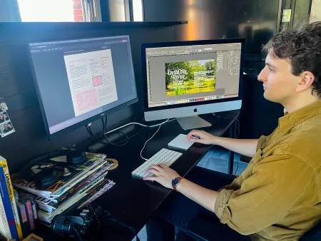 Evan Okun sits at a desk with two monitors, one showing a magazine rates page and the other an InDesign layout titled "Over the River and Down the Road."