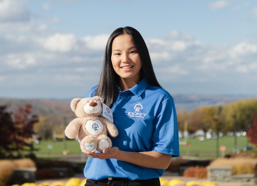 Adrienne Smith holds a teddy bear while standing outside with flowers behind her and Cayuga Lake in the distance.