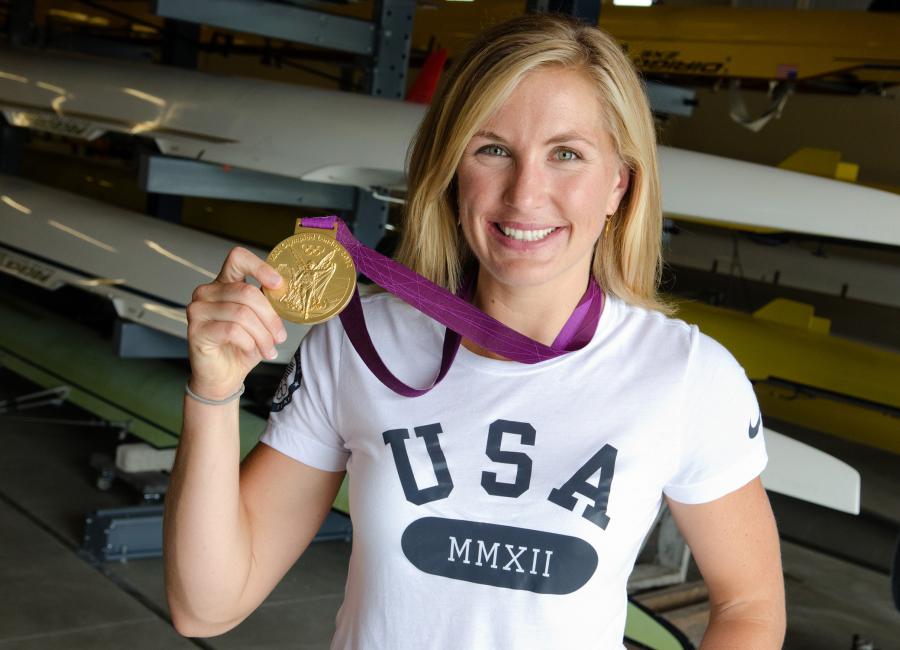 Meghan Musnicki holds up the Olympic gold medal around her neck while standing in a boathouse.