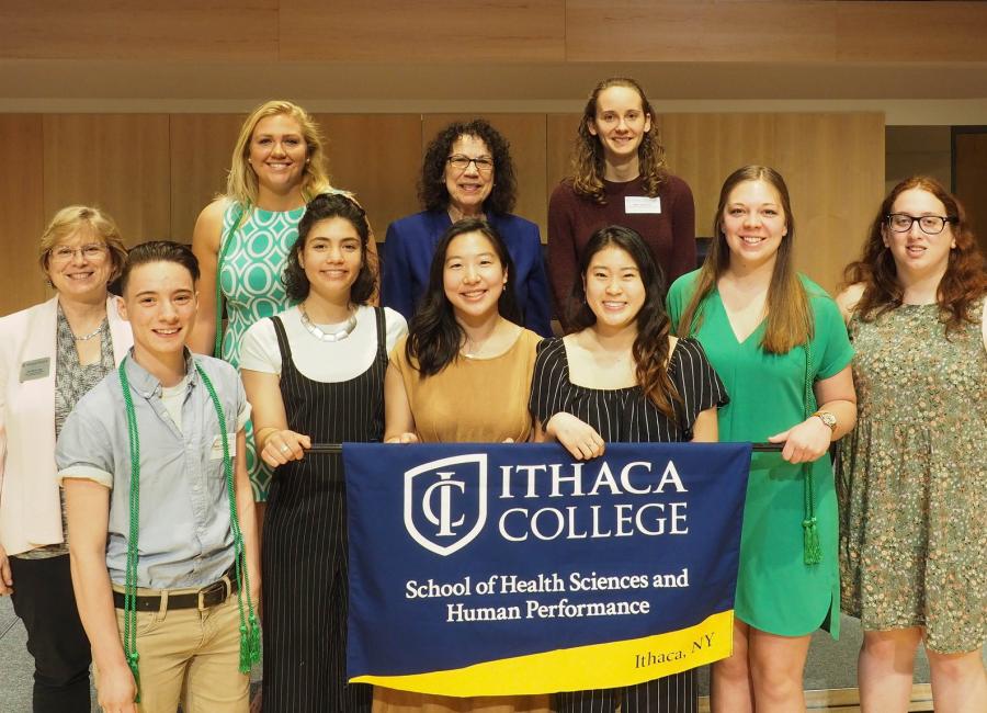 A group of students dressed nicely is standing on a stage holding a banner that says School of Health Sciences and Human Performance after an award ceremony.