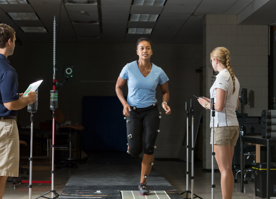 There is a student attached to sensors that are measuring her movement as she is running. There is a student standing to her left with a clipboard making notes and observations. as well as a student to the right doing the same thing.