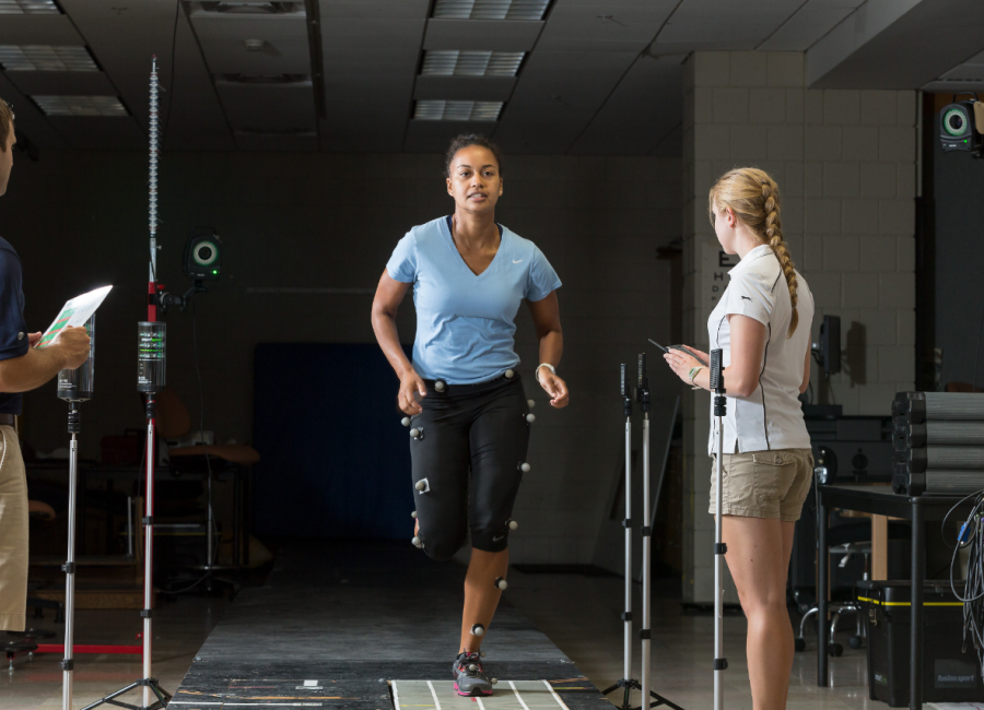 A student in a blue shirt is running on a sensor pad with sensors attached to her legs. Another student is standing nearby taking notes on a clipboard.