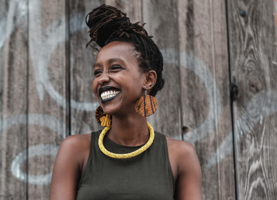A picture of a young, smiling Black woman.  Her hair is pulled into a front knot at the top of her forehead.  She is wearing a dark green tank top, dangling, round, wooden earrings and a yellow beaded necklace.  She is looking off to her right.  Behind her is a brown wooden fence with some white writing on it.