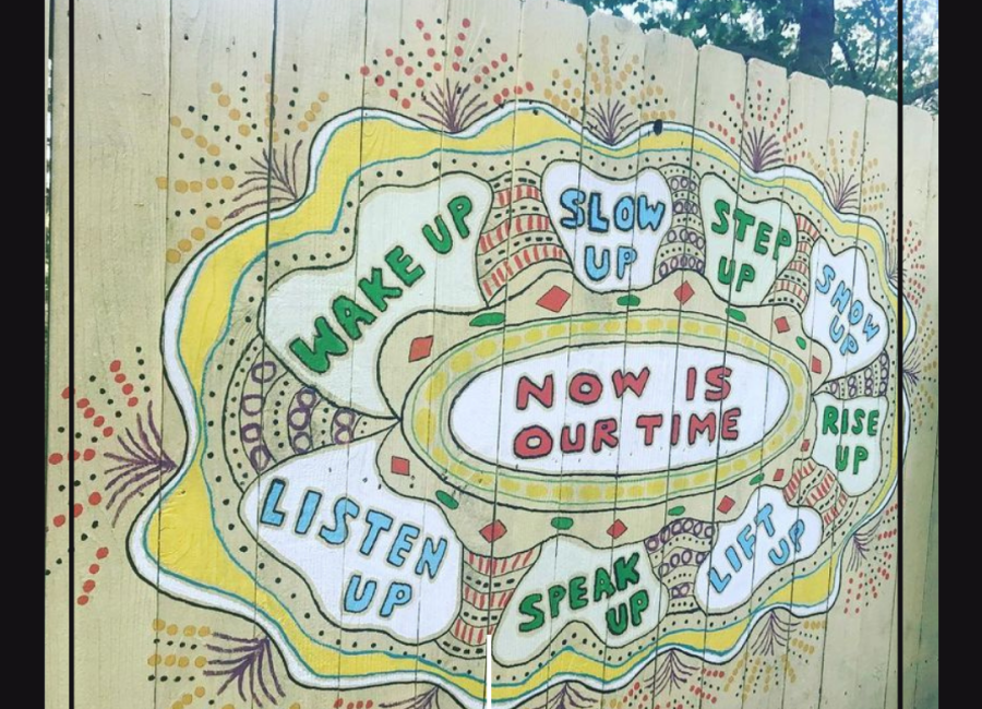 A mural painted on a light yellow fence.  The middle is a oval that reads "Now is our time" and is surrounded by bubbles that read, "wake up", "slow up", "step up", "show up", "rise up", "lift up", "speak up", and "listen up".  The outsides of the mural are decorated with sprays of dots and borders in white, yellow, and red.