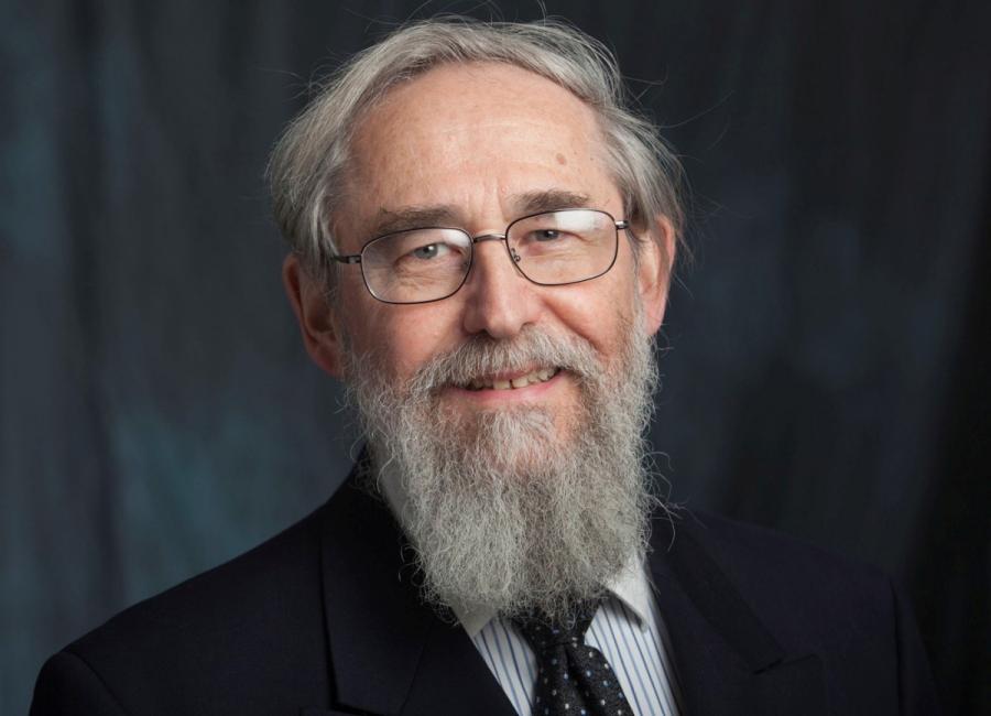 A picture of an older White man smiling at the camera.  He is wearing glasses and has a medium length grey beard that matches his grey hair.  He is wearing a dark suit and sits in front of a dark blue drop cloth.