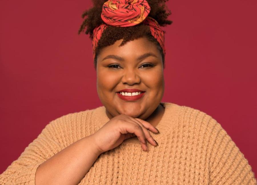 A picture of a young Black woman smiling directly at the camera.  She has a red and yellow cloth tied in her curly hair and is wearing a deep berry red lipstick.  She has her right hand placed beneath her chin and is wearing a wheat colored sweater with a waffle stitch pattern.  The background is a also a deep berry red color to match her makeup.