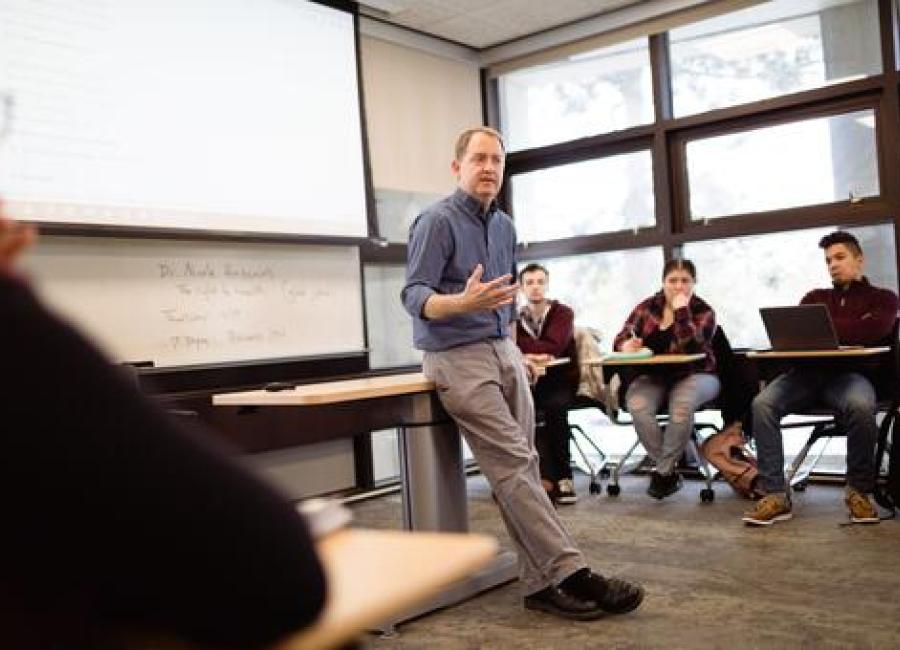 A man stands against a table in a classroom, talking to students