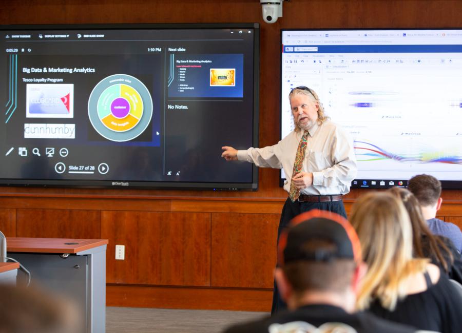 professor gestures to a display at the front of the classroom