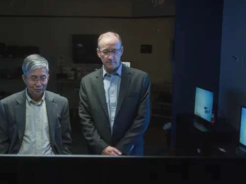 Chin Wan Tang and Steven Van Slyke stand in a darkened room surrounded by OLED monitors.