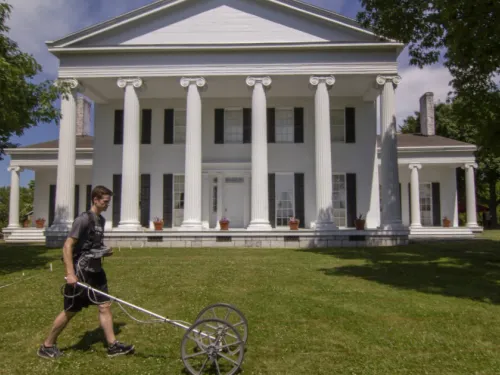 A man pushes a rolling machine in front of a white, Greek Revival style mansion