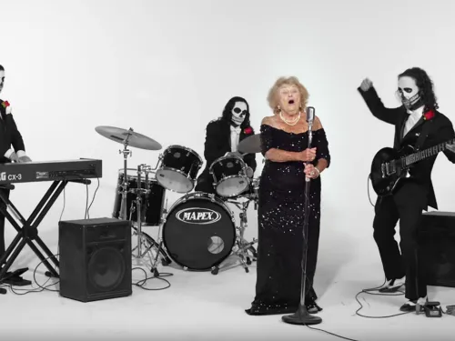 An older woman in black sings while men in black suits and skull makeup play instruments.