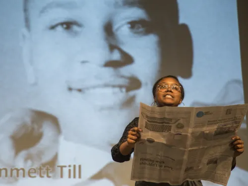 A young black woman in the foreground holds a newspaper while a photo of Emmett Till is projected on screen behind her.