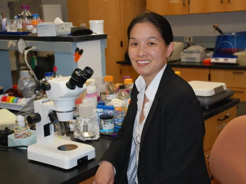 A woman sits and poses at a lab table, surrounded by scientific equipment. 