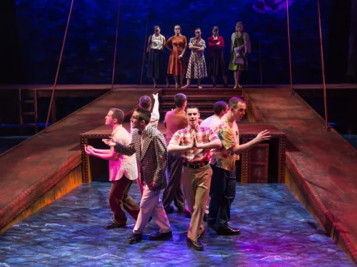 A group of men sing and dance on stage while women pose behind them