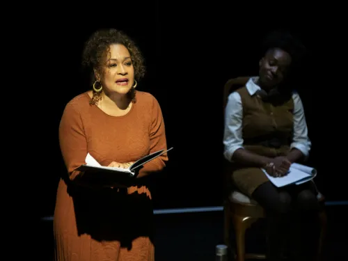 A woman in a spotlight reads aloud while another watches
