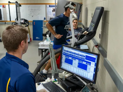 A man runs on a treadmill with a mask on as two others watch