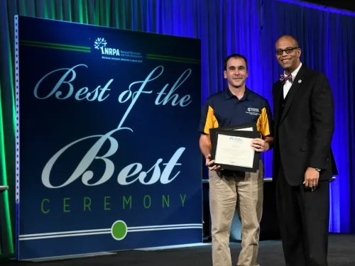 Two men pose with a certificate in front of a banner reading "Best of the Best"