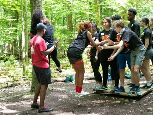 A young woman balances on ropes while other people help her balance