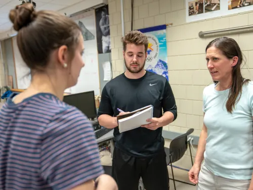 Two students are taking notes while speaking with a faculty member.