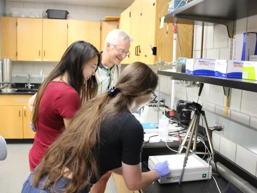 two women and their professor look at biomechanical equipment on a lab bench