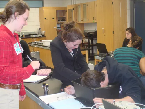 a student with a stethoscope monitors a fellow student whose face is down in a bucket on a lab bench, while another student records in a notebook