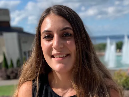This is a photo of Chasse Guerrera. Chasse is seated in front of the Ithaca College fountains wearing a black sleeve dress.