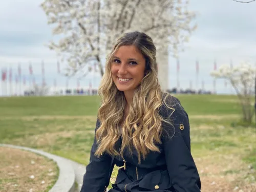 This is a photo of Danielle Pasquale. Danielle is seated outside, with green grass and a tree behind where Danielle is sitting. Danielle is wearing a black coat and pants.