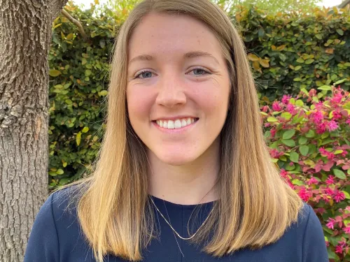 This is a photo of Hannah Thomas. Hannah is smiling at the camera with long blond hair. Hannah is wearing a long sleeve blue shirt. Hannah is standing outdoors, in front of a tree.