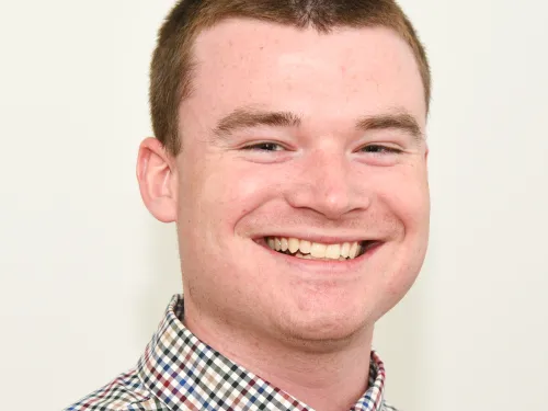 This is a photo of Jonathan Ripic. Jonathan is wearing a checkered shirt and is smiling at the camera. Jonathan has short hair, cut close.