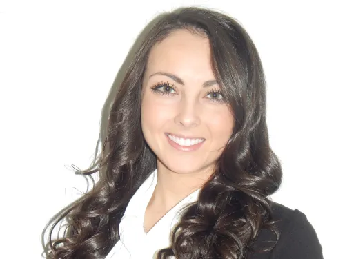 This is a photo of Natalie Knight. Natalie is wearing a white ruffled shirt underneath a black blazer. Natalie has long dark curly hair.