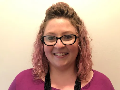 This is a photo of Sarah O'Hagen. Sarah is wearing a long sleeve purple shirt and a long black necklace around the neck. Sarah is wearing glasses.