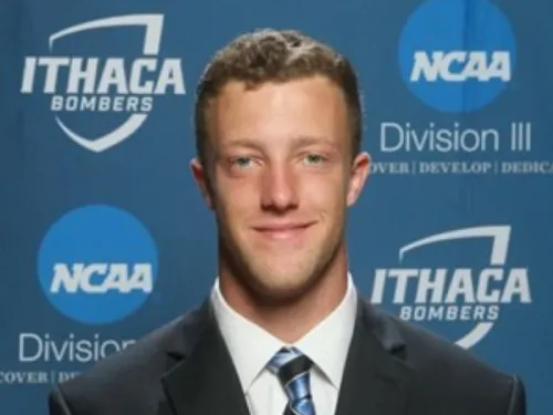 This is a photo of Tyler Winslow. Tyler is wearing a dark blue suit with a white shirt and blue striped tie. Tyler is standing in front of an Ithaca Athletics Division III background.