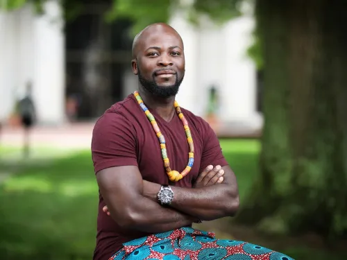 a young man sits with his arms folded in a redish shirt, necklace, and closed smile.