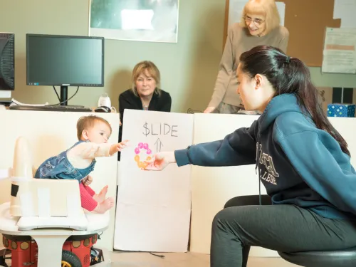 A baby in a small robotic chair alongside a college student reaching their hand out to entice the baby to move.
