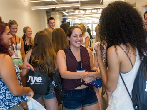 A student in a maroon tank top is shaking the hand of a new student to campus welcoming them.