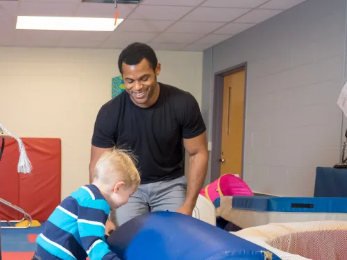A man in a black shirt is watching a young child play with gym equipment.