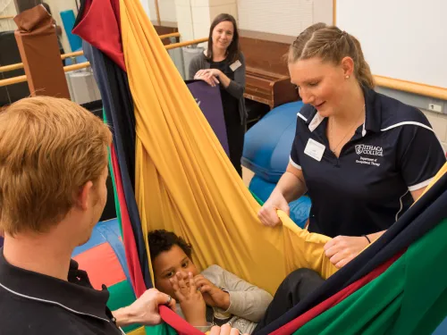 Two students are looking into a colorful hammock where a child is swinging.