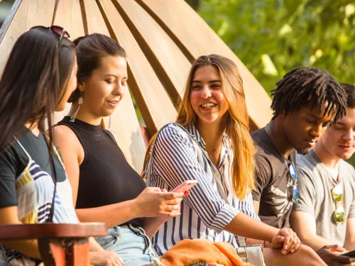 A group of 5 students are seated together on a bench smiling and laughing.
