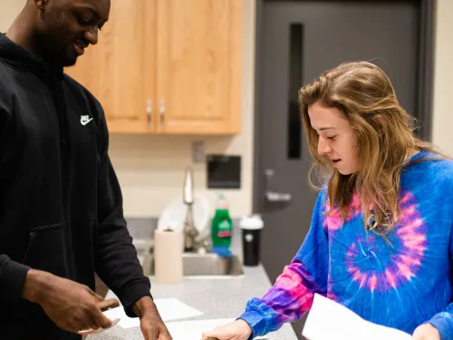 A student in a black shirt is cutting up food on a cutting board while a student in a tie-dyed shirt looks on.