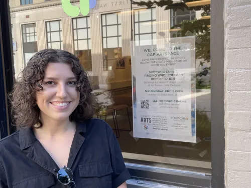 Photo of young woman standing in front of a large window outdoors. Window has community arts partnership logo decal.