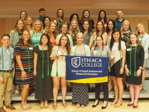 A group of about 30 students are standing on stage holding a banner for the school and have green cords draped around their necks to represent induction into the Alpha Eta Honor Society.